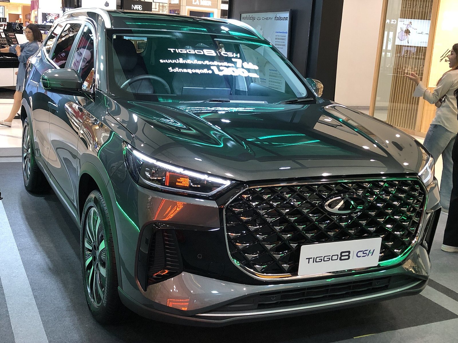 Σκουροπράσινο Chery Tiggo 8 CSH με μεγάλη μάσκα με μοτίβο διαμαντιού, σε μπροστινή τριτέταρτη όψη, εκτεθειμένο μέσα σε εμπορικό κέντρο