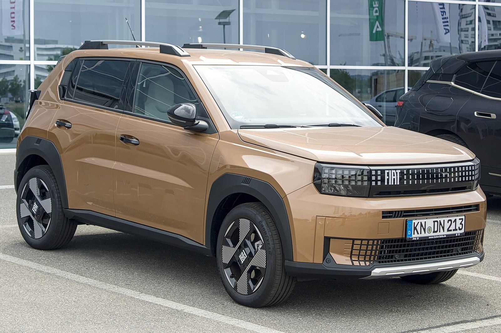 Χάλκινο Fiat Grande Panda Hybrid με μαύρα προστατευτικά θόλων και μπάρες οροφής, σε μπροστινή τριτέταρτη όψη, σταθμευμένο έξω από έκθεση αυτοκινήτων