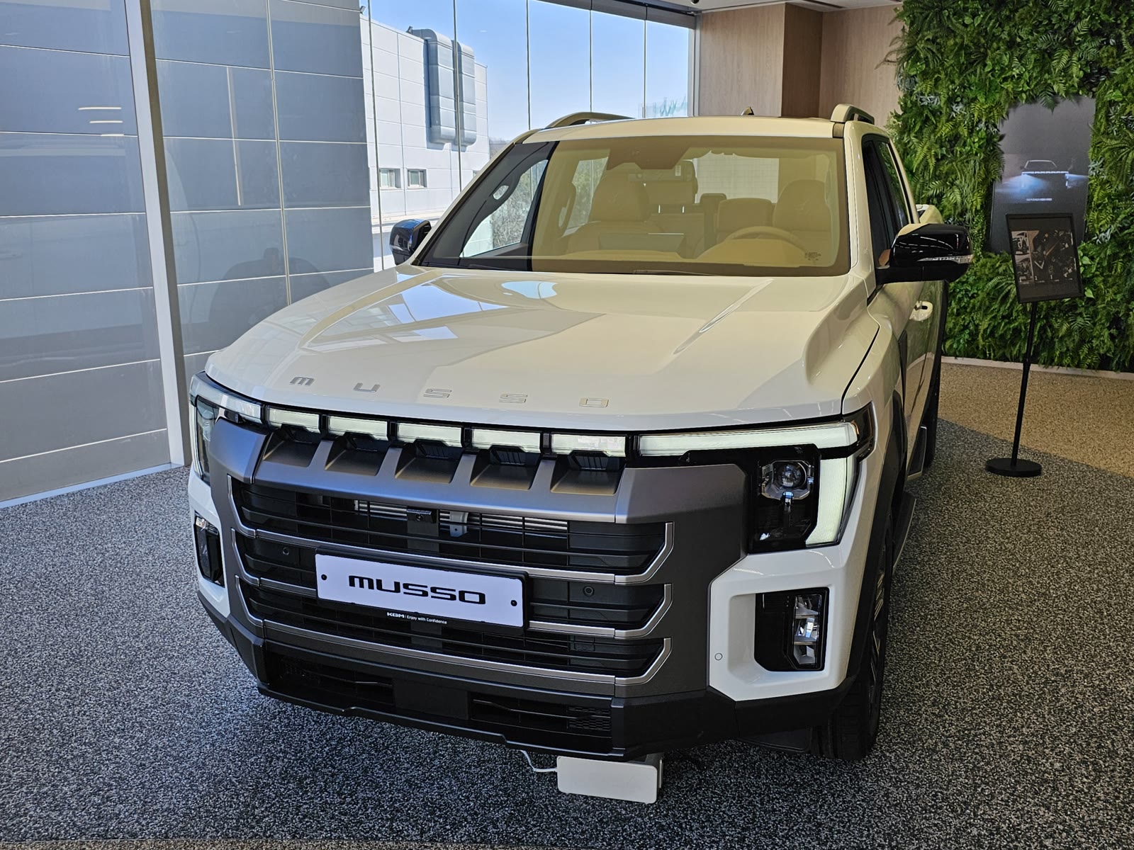 Λευκό KGM Musso Grand 2.2 diesel με μακριά καρότσα, σε μπροστινή τριτέταρτη όψη, μέσα σε εκθεσιακό χώρο